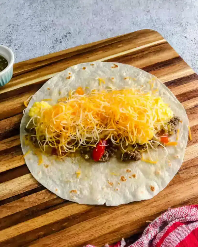 Breakfast burrito assembly—tortilla topped with eggs, sausage, peppers, and shredded cheese on a cutting board