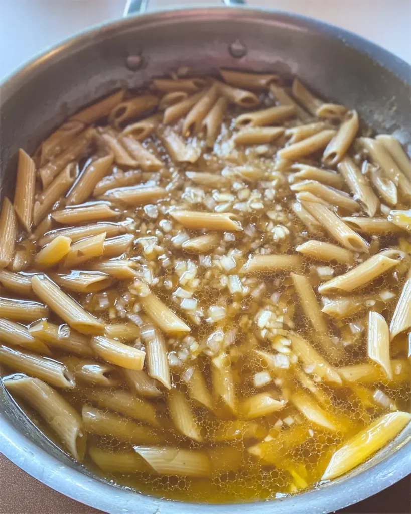 Creamy Garlic Parmesan Chicken Pasta 3 Penne pasta simmering in chicken broth with onions in a stainless steel skillet